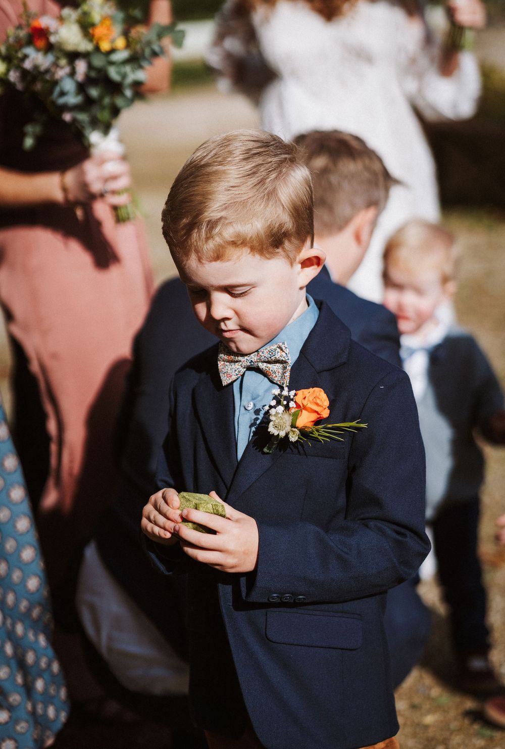Young ring bearer in navy suit checks his phone during an outdoor wedding ceremony.