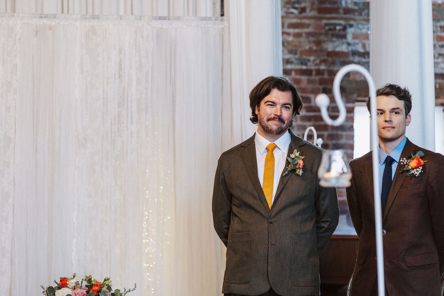 Two groomsmen in formal attire stand at altar decorated with white drapery and floral arrangements.