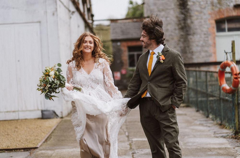 A joyful couple runs along an industrial walkway, with the bride in a flowing lace gown holding a bouquet.