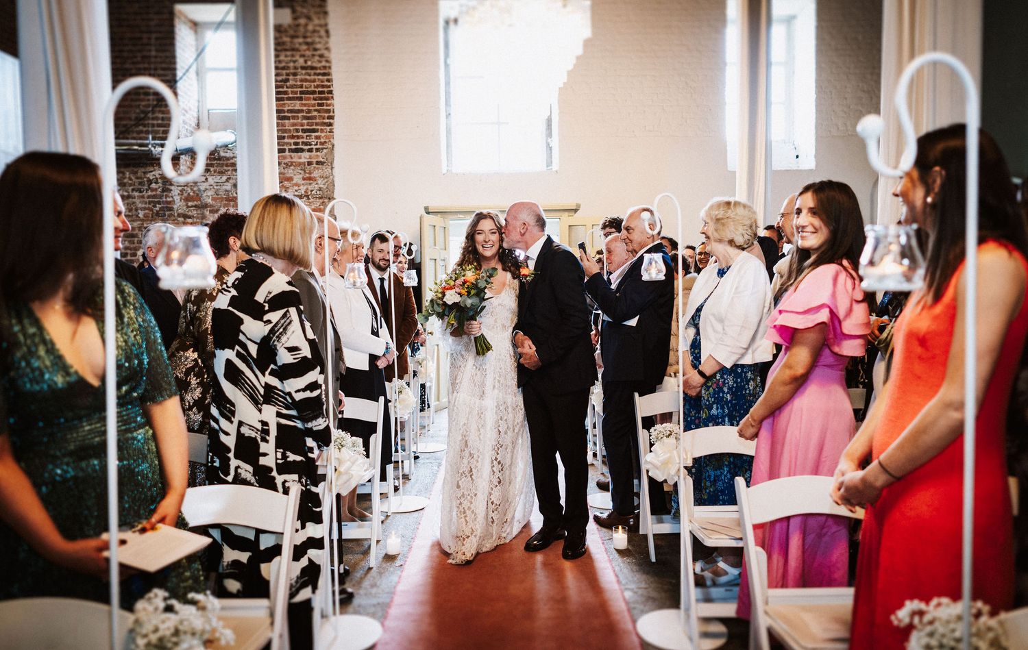 Wedding guests mingle and celebrate at an elegant indoor reception venue with exposed brick walls and natural lighting.