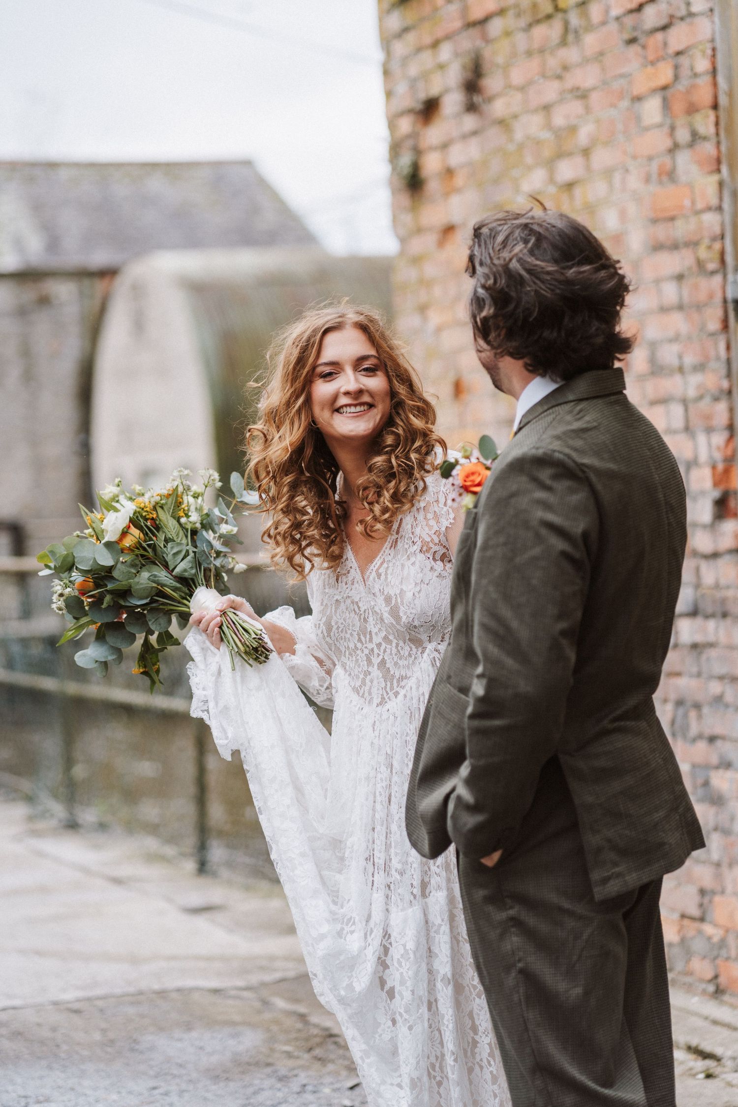 Romantic wedding scene with couple in white lace dress and dark suit embracing against rustic brick wall backdrop.