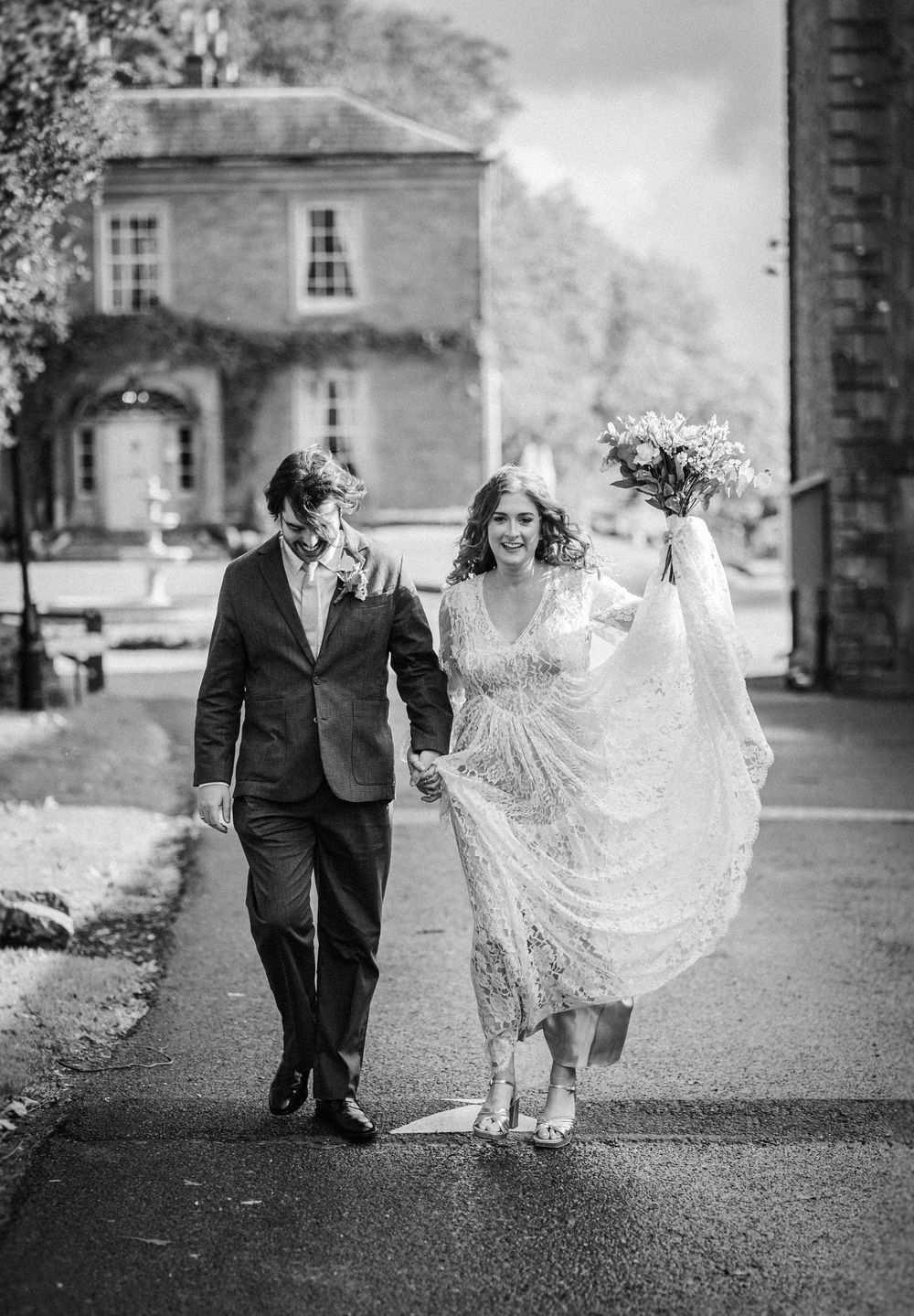 A couple walks together on a path in black and white, with the bride's lace dress flowing in the wind.