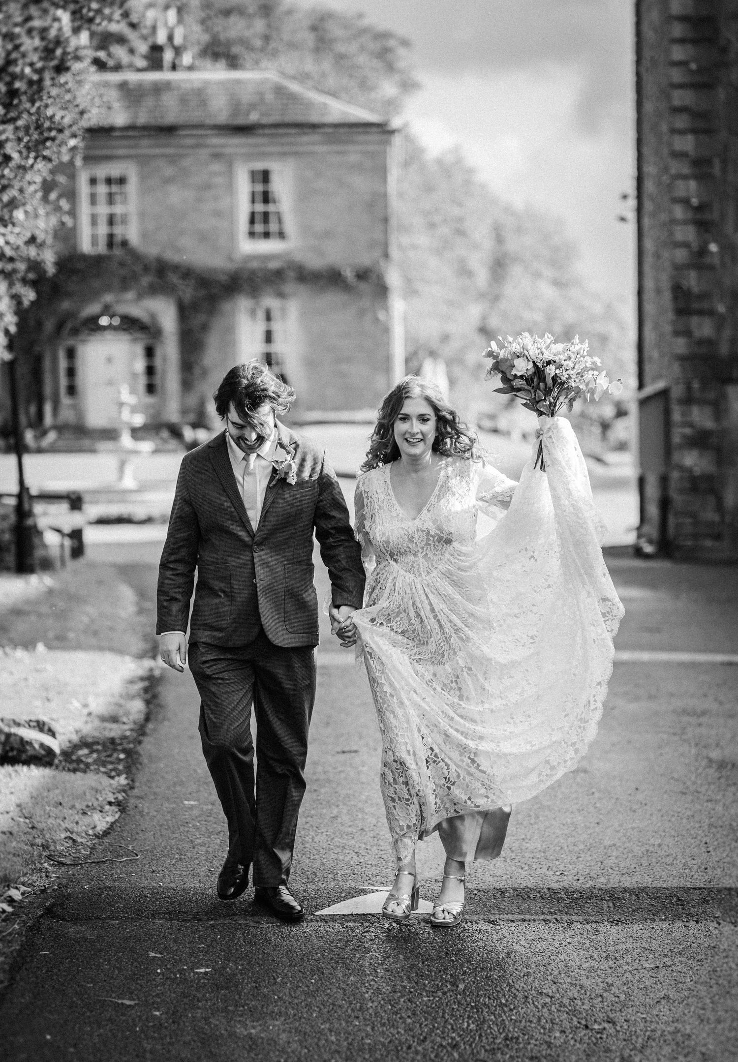 A couple walks together on a path in black and white, with the bride's lace dress flowing in the wind.