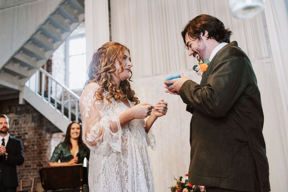 A couple exchanges rings during an intimate indoor wedding ceremony in a rustic brick-walled venue.