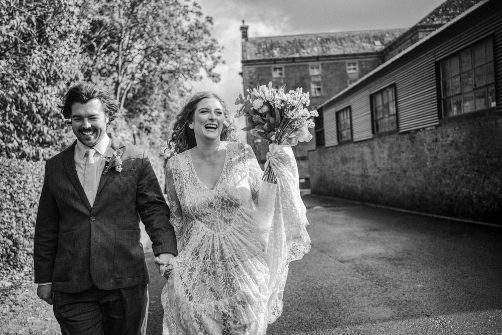 A joyful couple walks together outdoors on their wedding day, with the bride holding a bouquet in black and white.