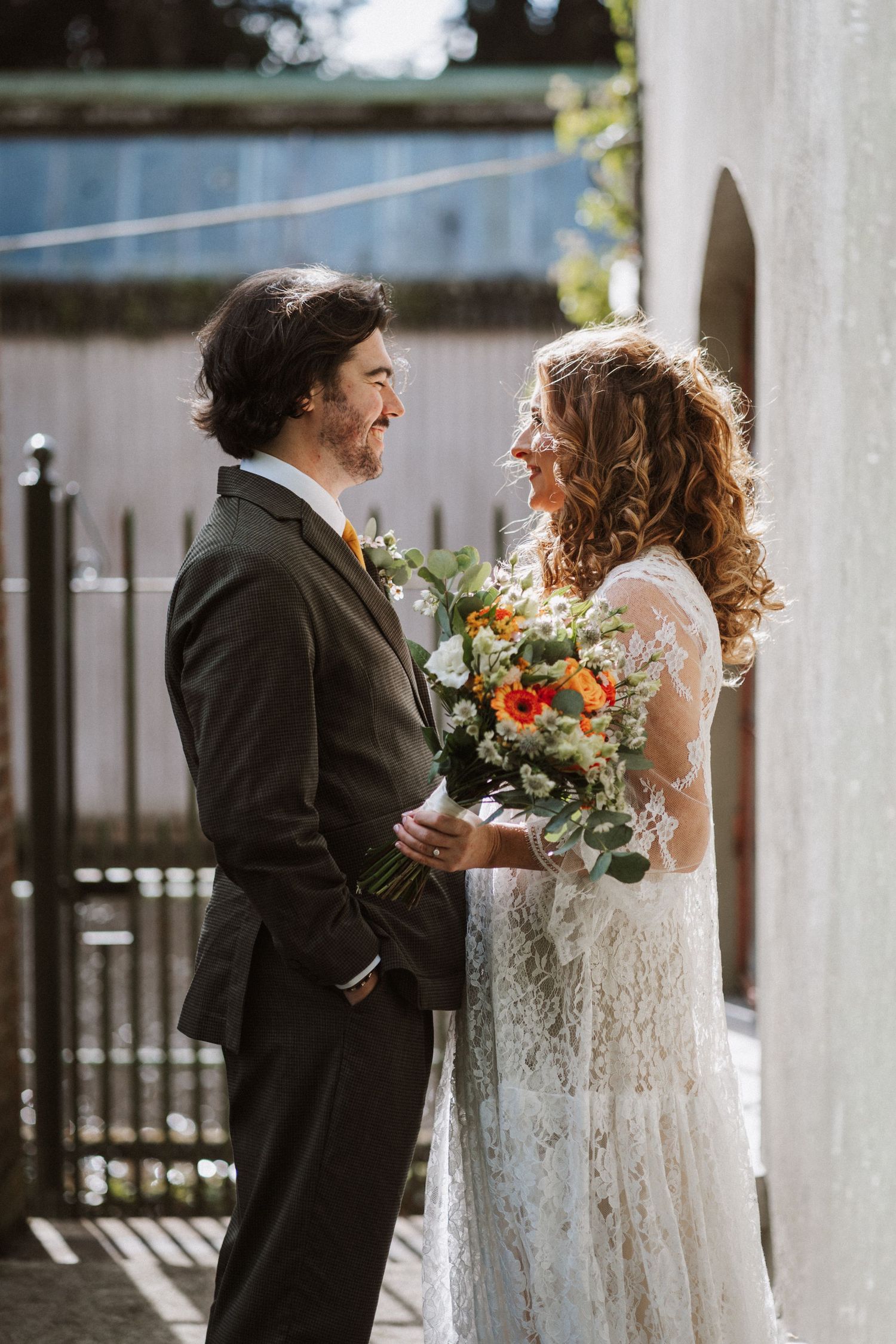 Bride in lace gown shares a tender first look moment with wedding guest in black suit on sunny outdoor patio.