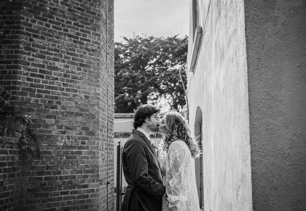 A black and white photo of a couple in wedding attire standing near a brick wall.