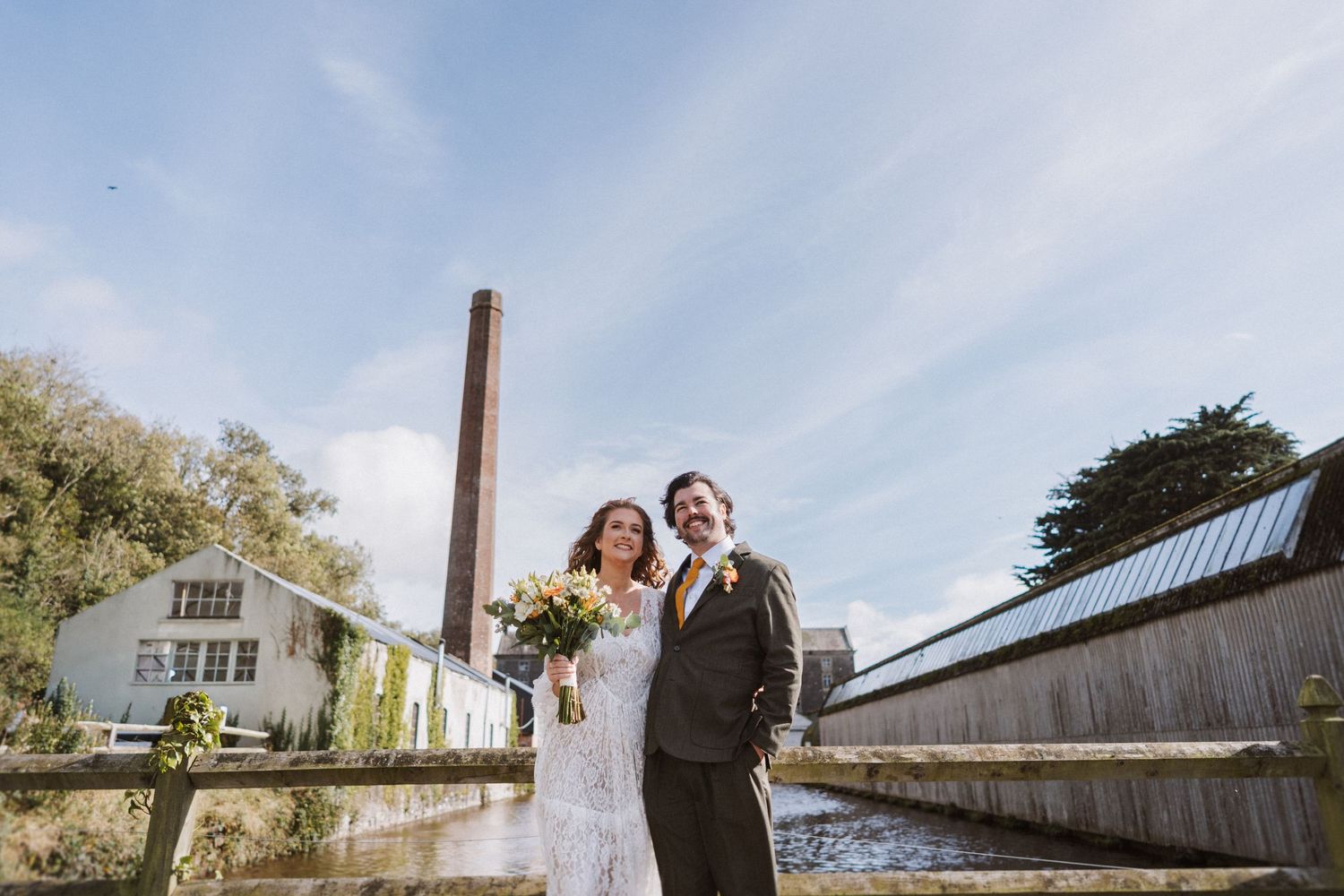 A couple poses together on their wedding day in front of rustic wooden buildings and autumn trees.