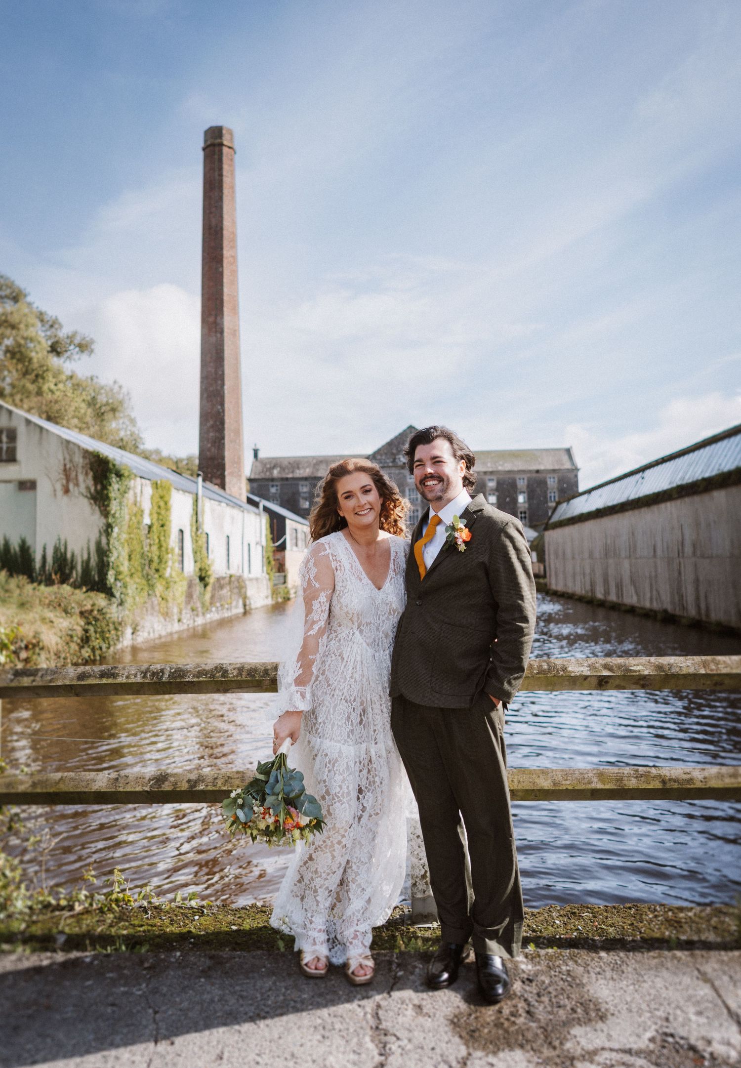 A couple in wedding attire poses playfully by a waterfront with industrial buildings and a tall brick chimney in the background.