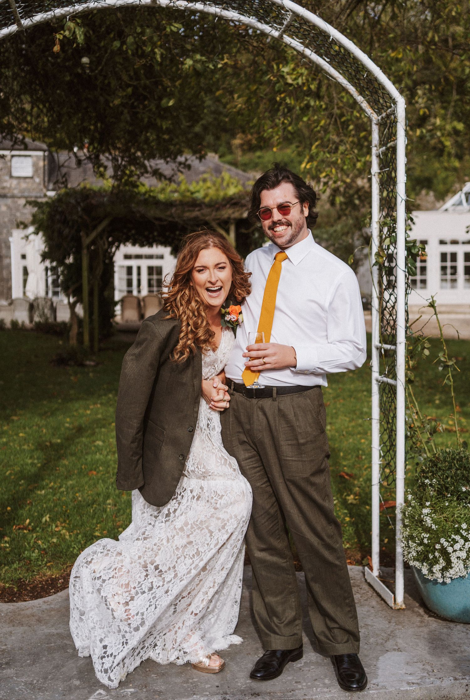Playful wedding couple toasts with beer bottles while sitting on a white metal bench in an outdoor garden setting.