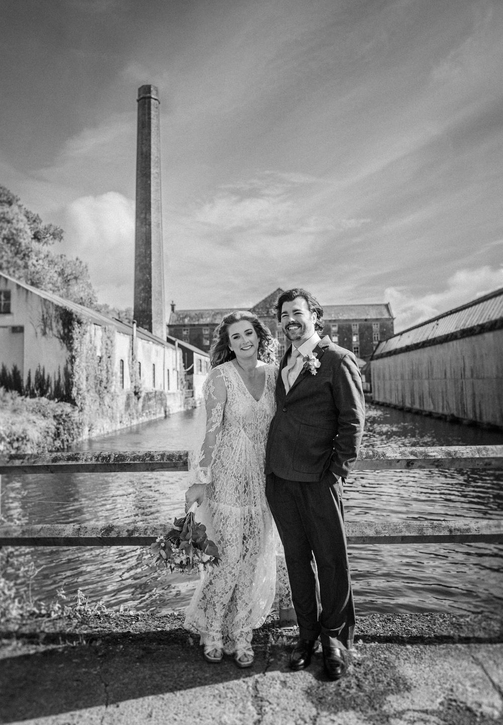 A couple poses playfully near a tall industrial smokestack by the water on an overcast day.