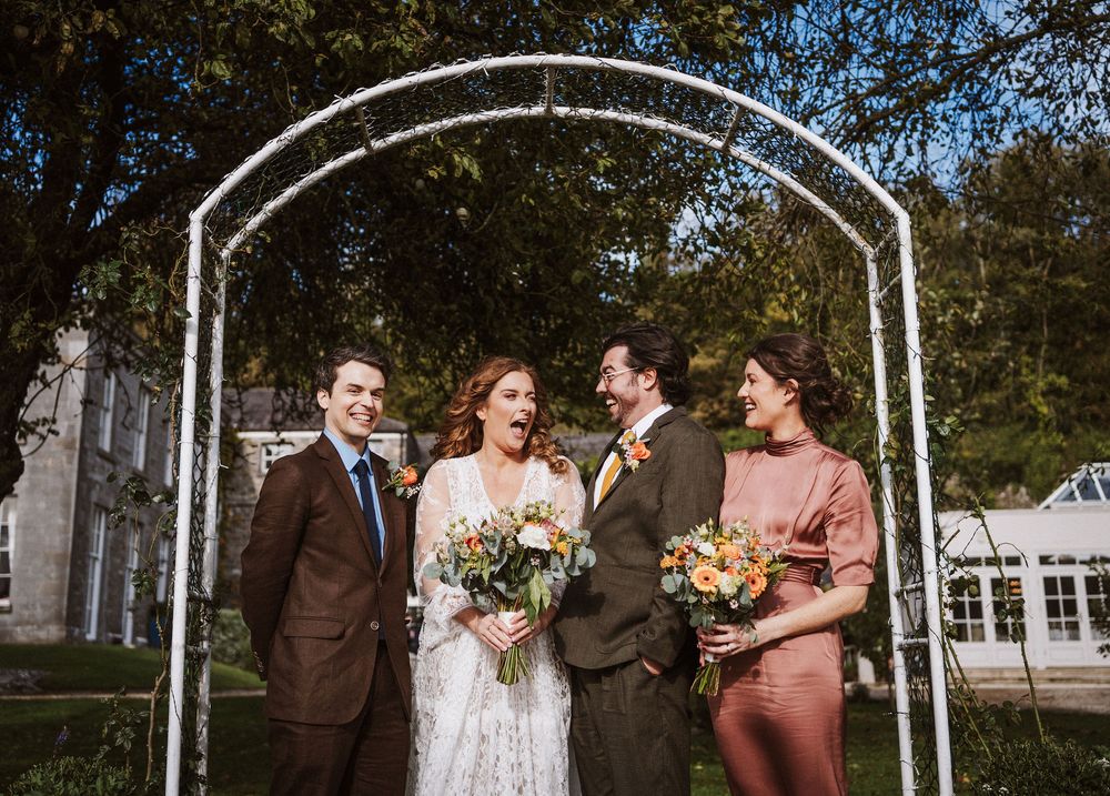 A wedding party stands together under a white arch during an outdoor ceremony with trees in the background.