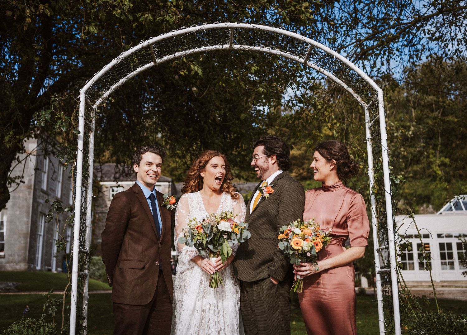 A wedding party stands together under a white arch during an outdoor ceremony with trees in the background.
