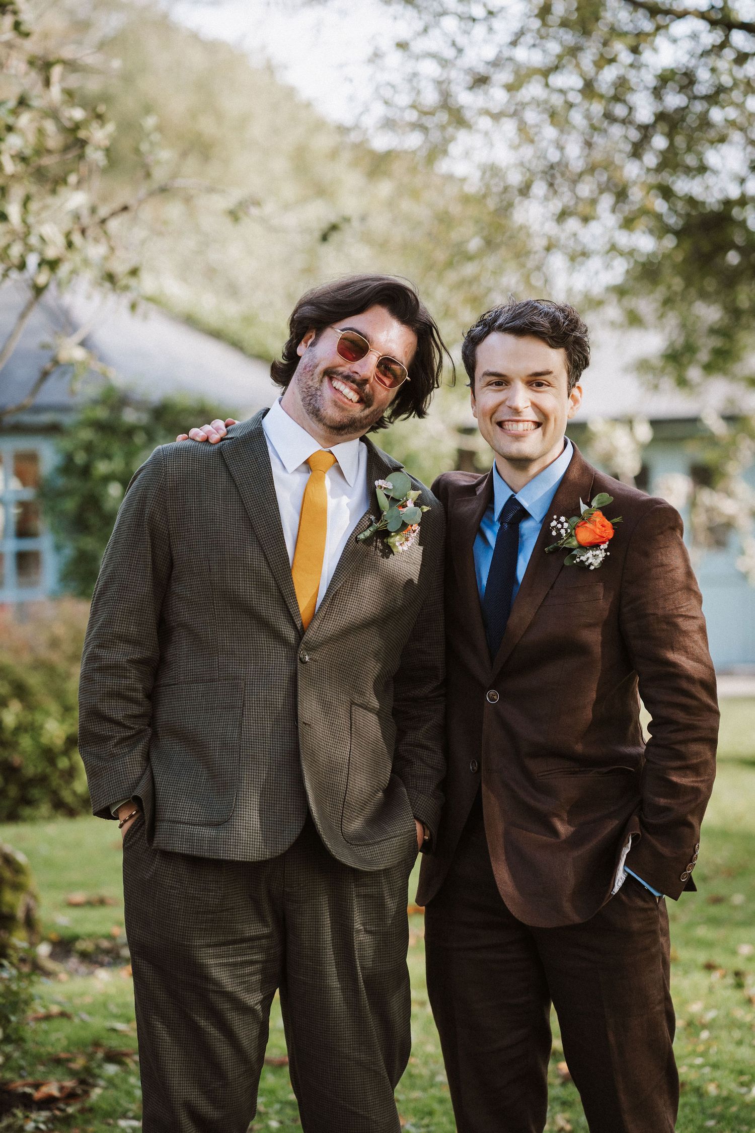 Two groomsmen in suits smile together at an outdoor wedding, wearing yellow and blue ties with boutonniere flowers.