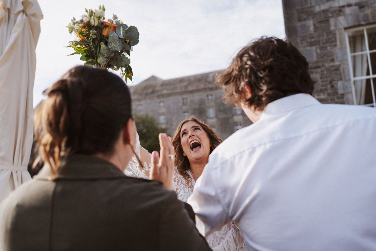 A group of wedding guests dancing and celebrating at an outdoor reception near a stone building.