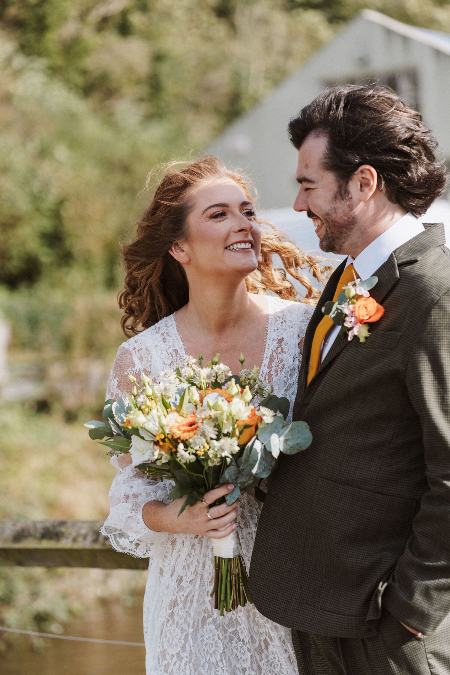 A couple shares a joyful moment on their wedding day, with the bride holding a vibrant orange and white bouquet.