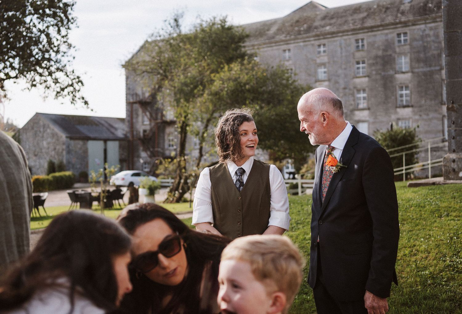 Wedding guests mingle and socialize on a grassy lawn outside a grand stone building on a cloudy day.