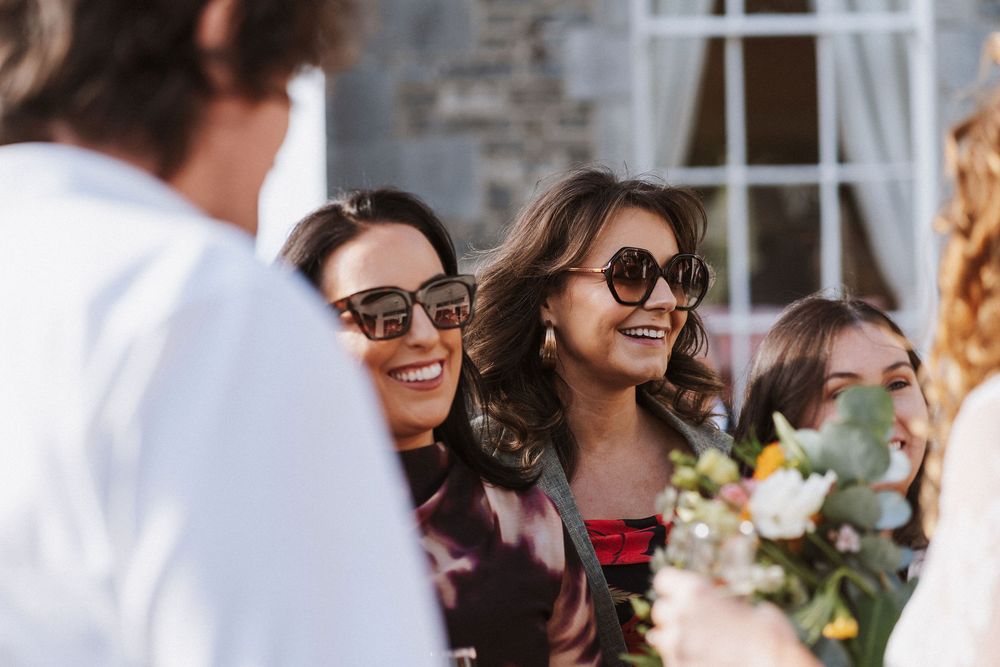 Guests at an outdoor wedding ceremony smile and chat while seated in white chairs surrounded by natural decor.