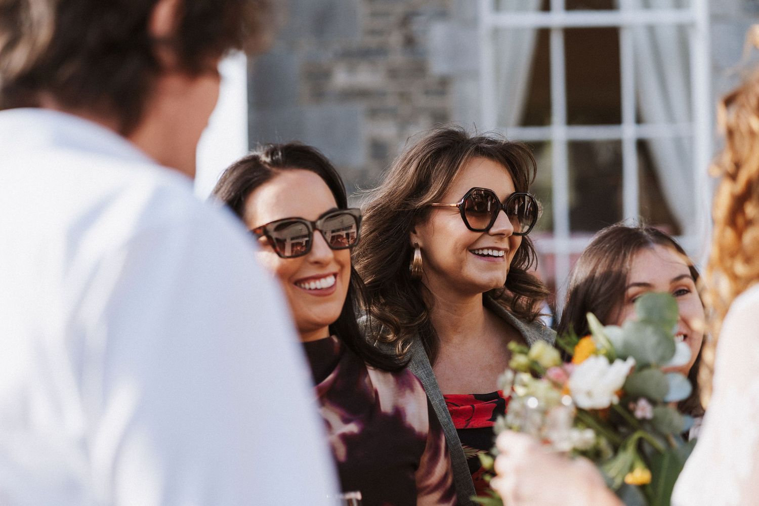 Guests at an outdoor wedding ceremony smile and chat while seated in white chairs surrounded by natural decor.