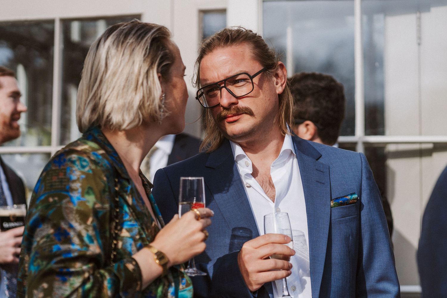 Two people in formal attire having an animated conversation while holding champagne glasses at an outdoor event.