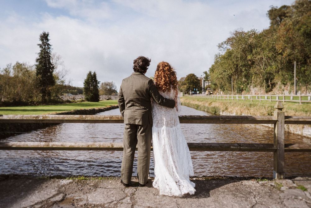 A couple walks hand in hand along a scenic lakeside path during an autumn wedding photoshoot.