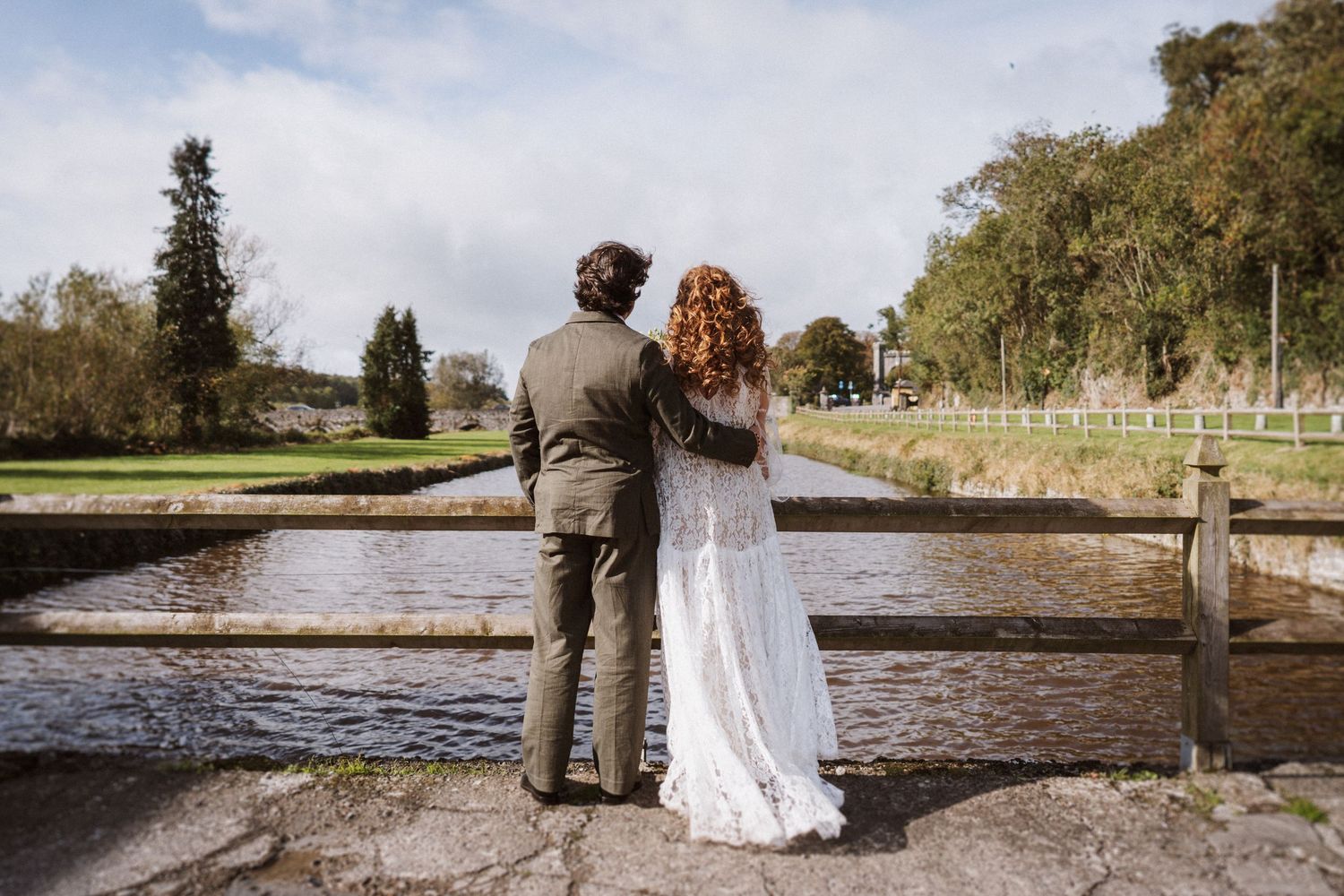 A couple walks hand in hand along a scenic lakeside path during an autumn wedding photoshoot.