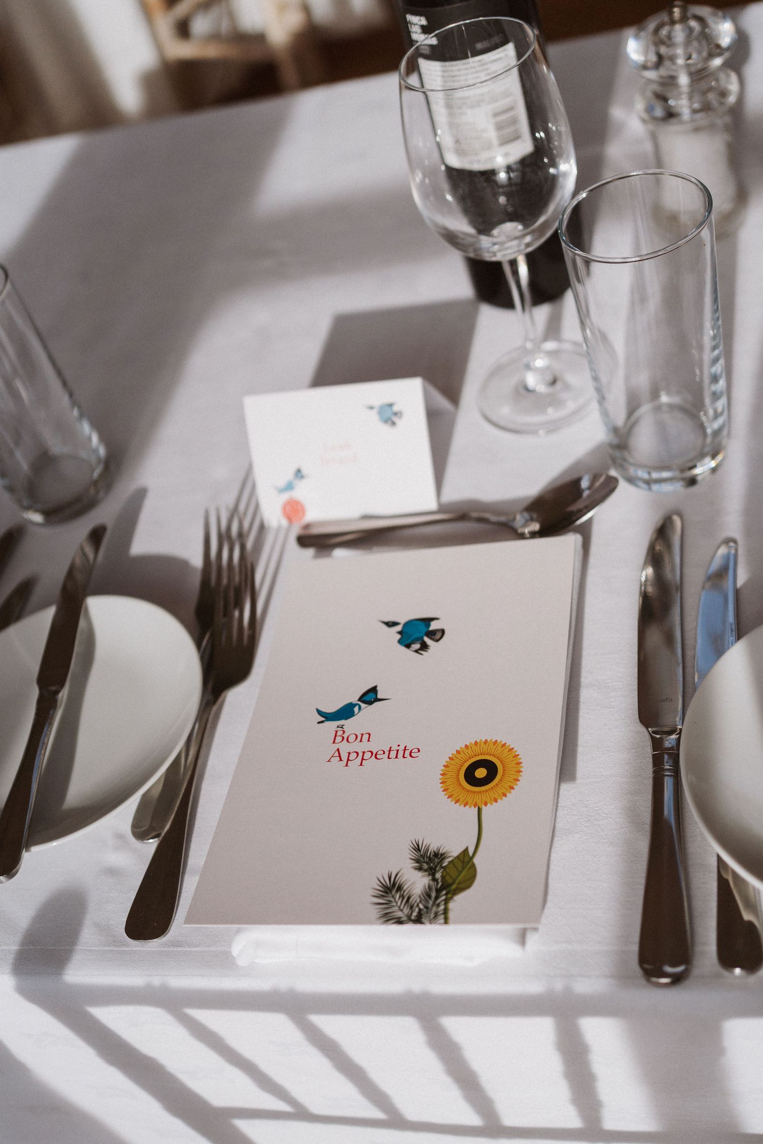 Restaurant table setting with wine glasses and menu decorated with blue birds and sunflower illustration on white tablecloth.