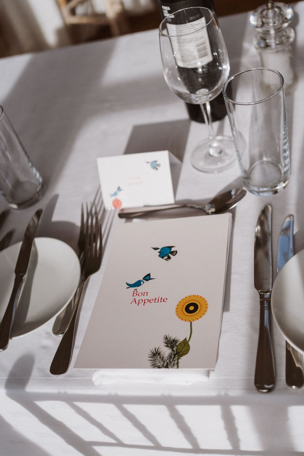 Restaurant table setting with wine glasses and menu decorated with blue birds and sunflower illustration on white tablecloth.