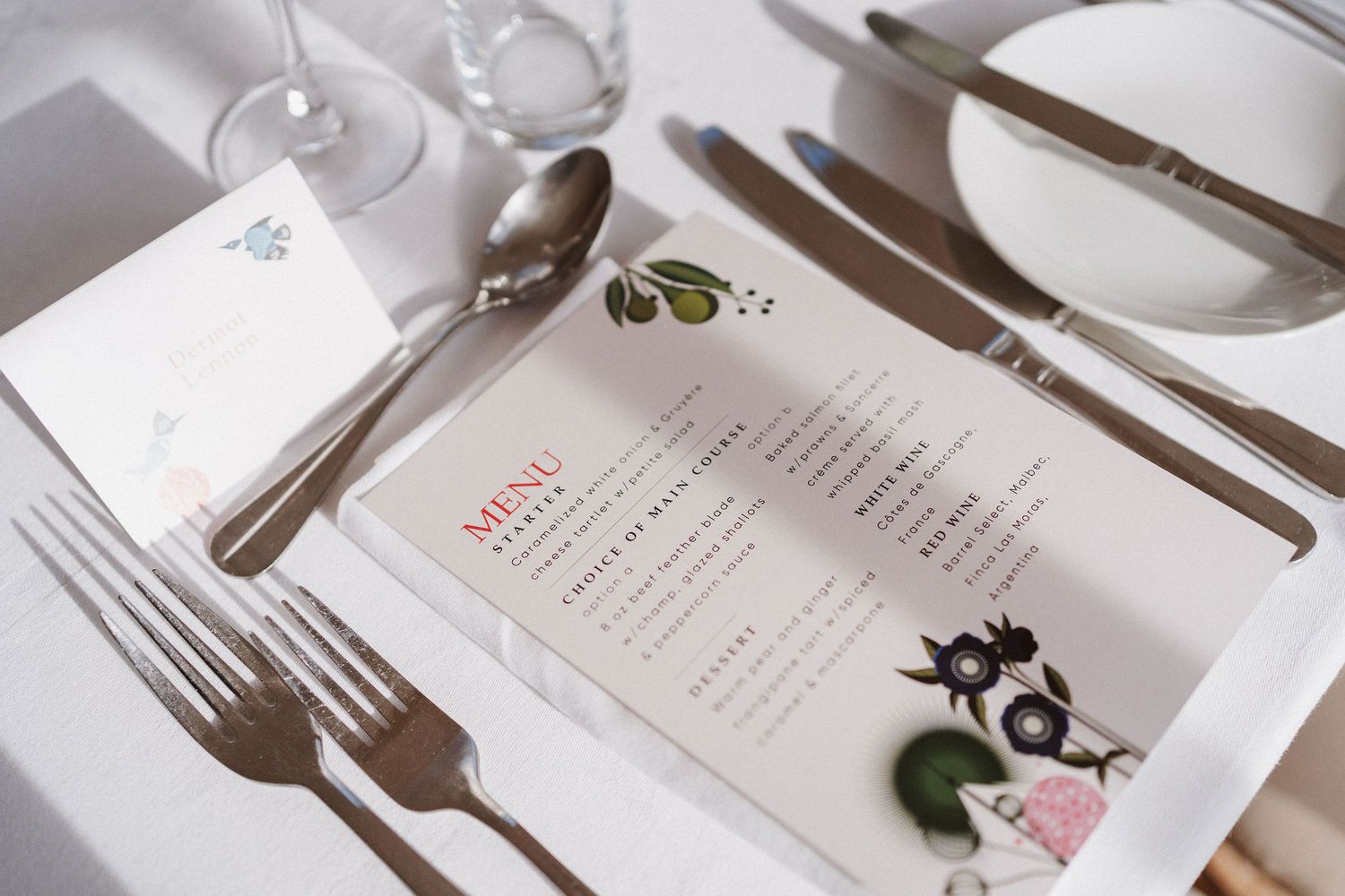 Elegant table setting with white plates, silver cutlery, and printed menus arranged on a light colored surface.