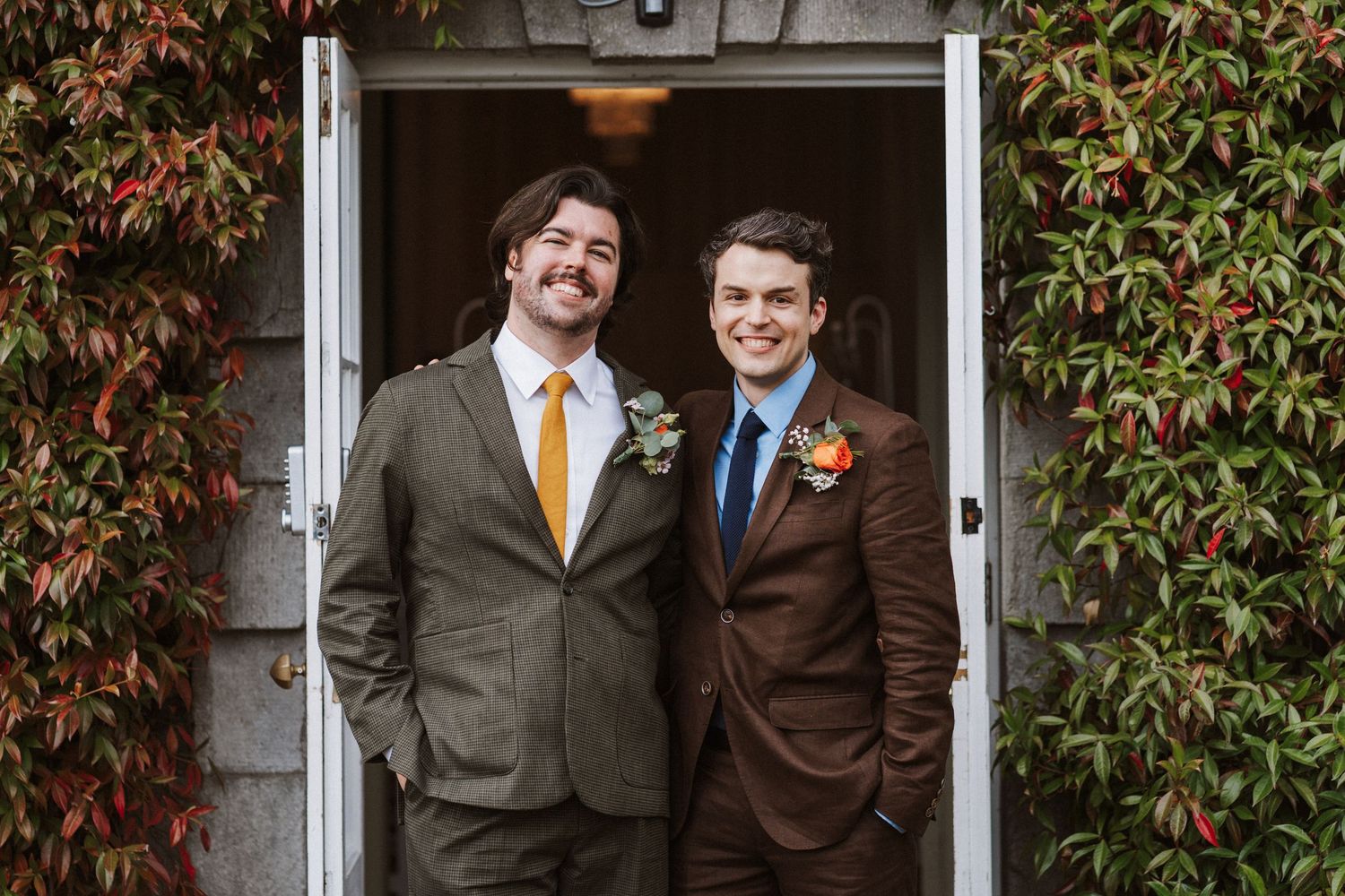 Two people in suits pose together against a brick wall with autumn colored ivy leaves.
