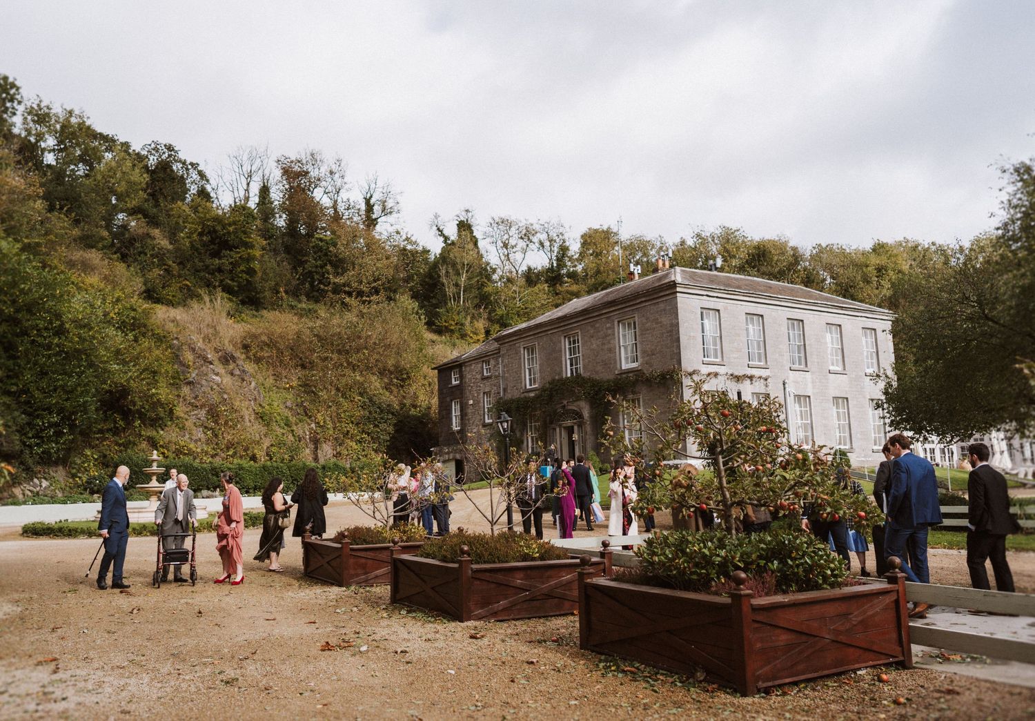 Wedding guests gather outdoors for a celebration at a rustic venue with historic buildings and natural landscaping.