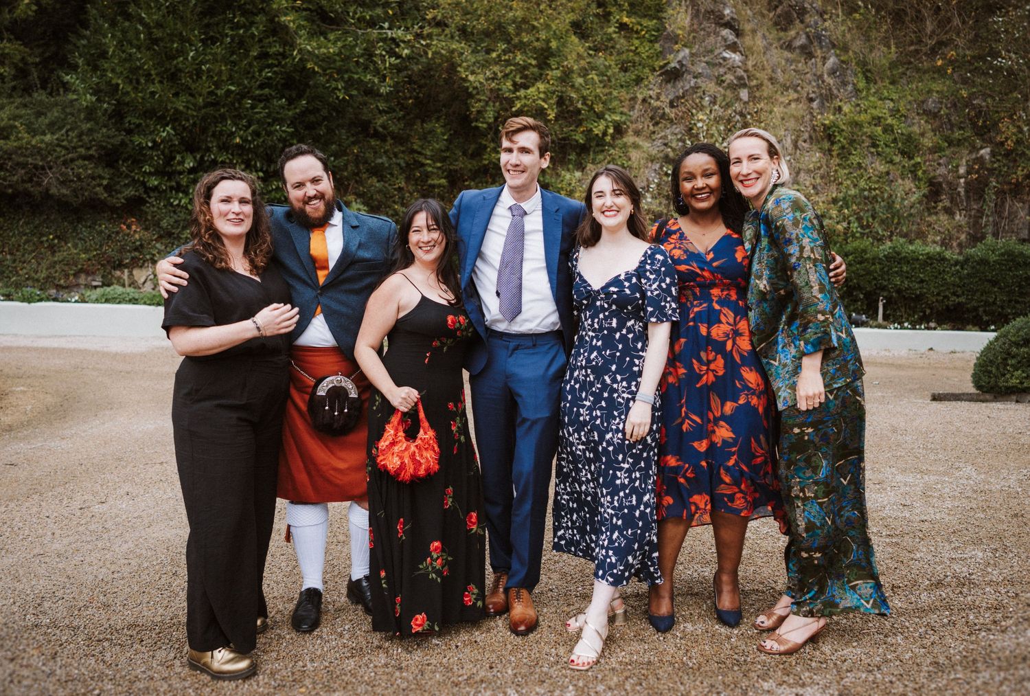 A group of wedding guests in formal attire pose together outdoors in a natural setting during a fall celebration.