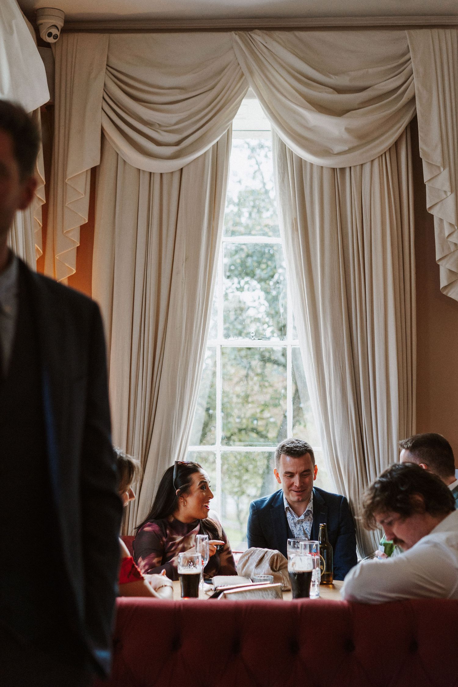 Elegant dining room scene with draped cream curtains and guests seated at tables in warm lighting.
