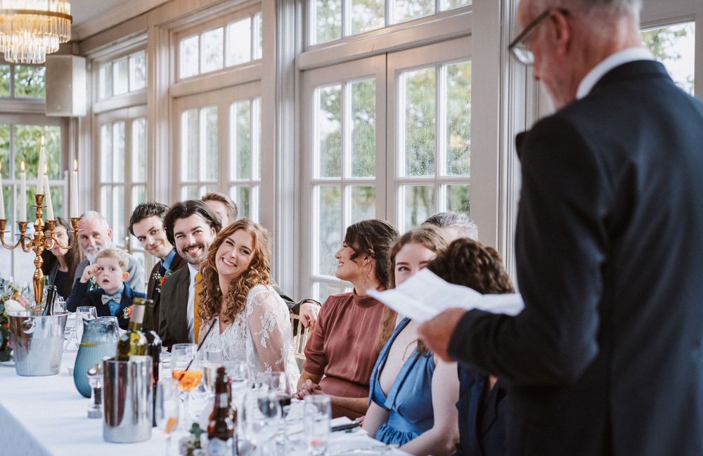 Wedding guests seated at elegant tables beneath a crystal chandelier in a bright, window-lined reception venue.