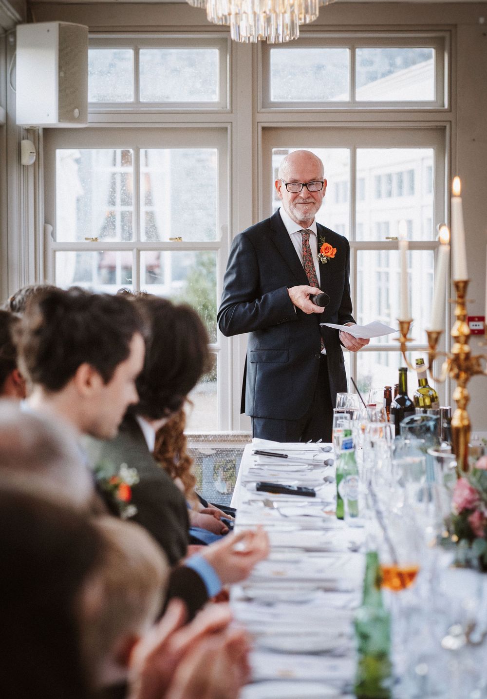 A speaker delivers a wedding toast at an elegant reception dinner with guests seated at long tables.