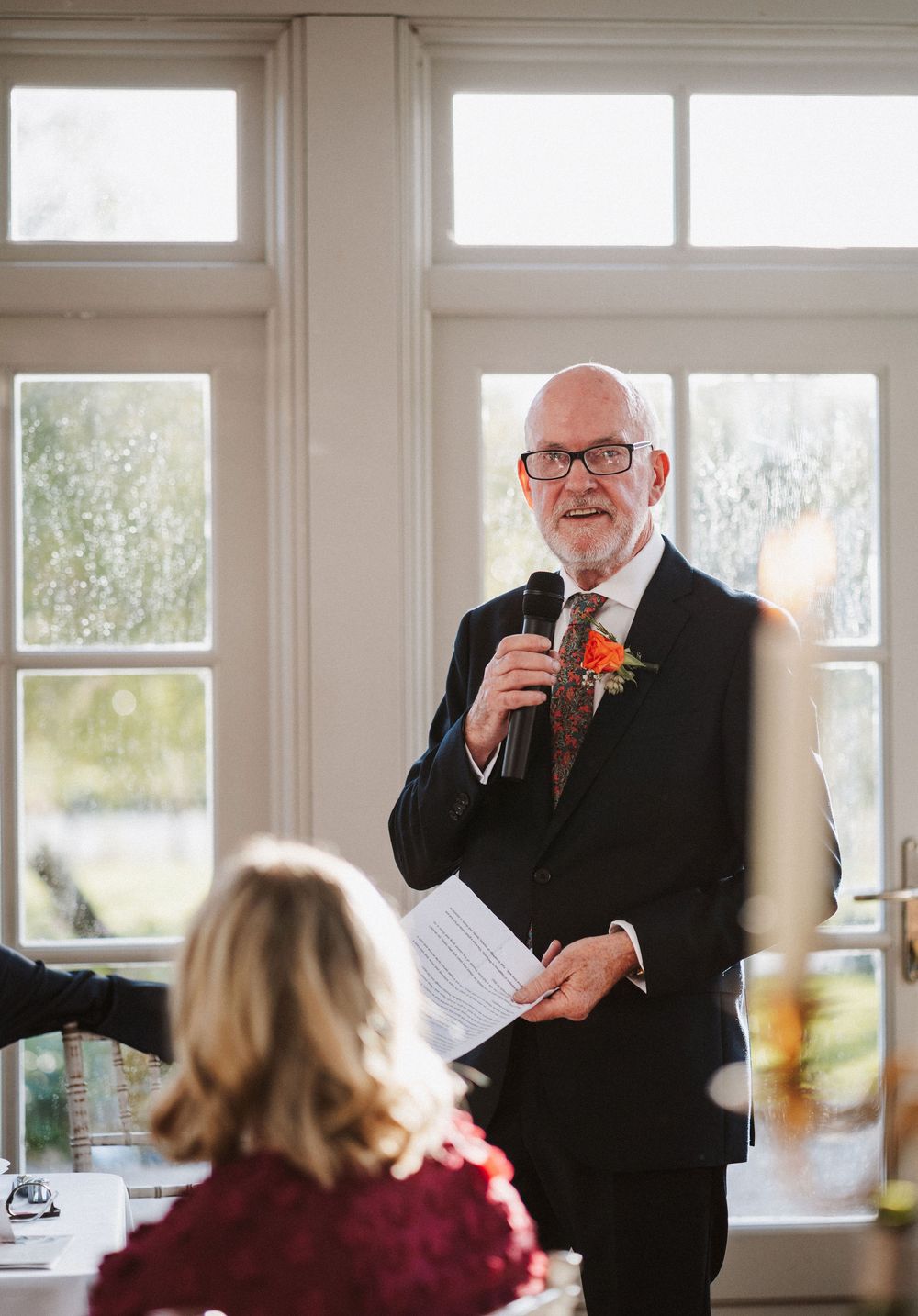 A speaker in a black suit with orange boutonniere addresses guests at an elegant indoor venue with large windows.