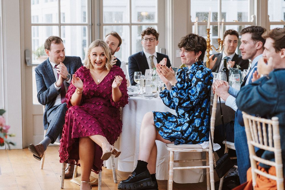 Wedding guests laugh and applaud while seated at elegant reception tables in a bright venue with large windows.