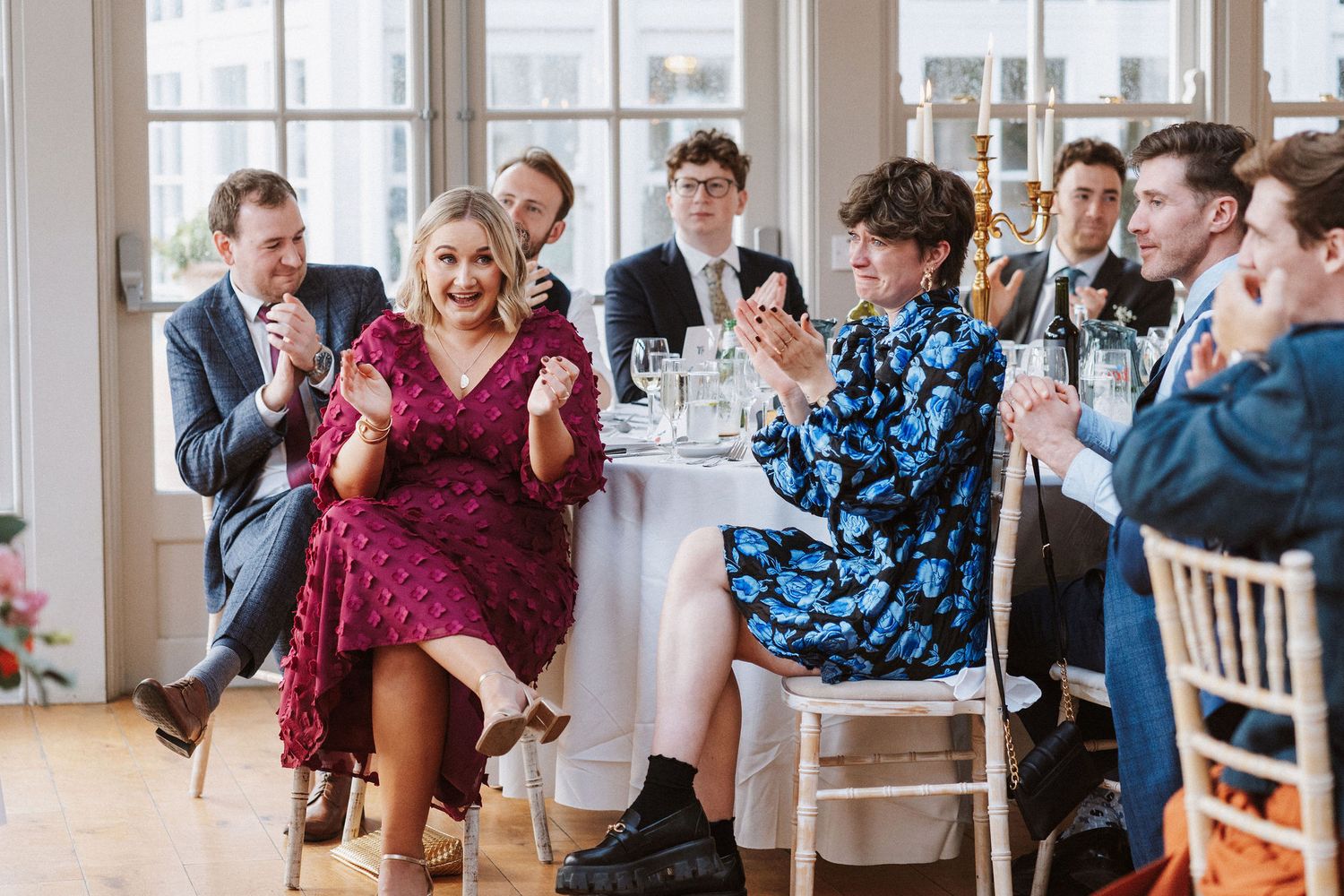 Wedding guests laugh and applaud while seated at elegant reception tables in a bright venue with large windows.