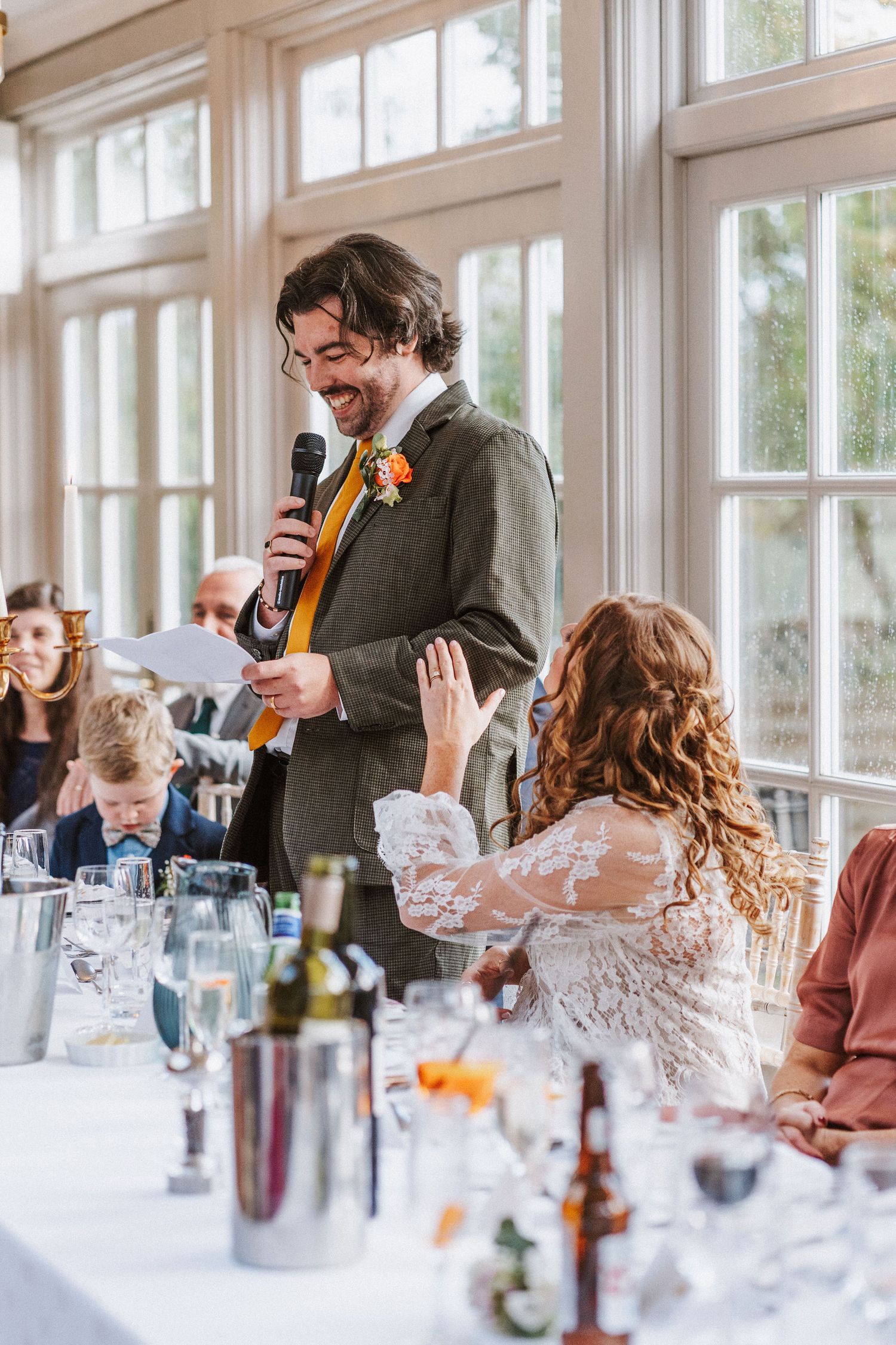 A wedding reception speech at a bright sunlit venue with guests seated at a decorated table with drinks and flowers.