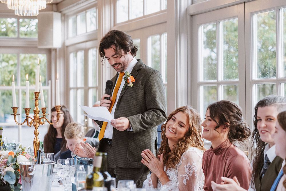 Wedding guests at a reception share joyful laughter while seated at elegant tables in a bright sunlit venue.