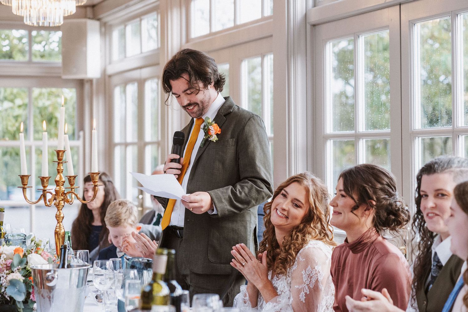 Wedding guests at a reception share joyful laughter while seated at elegant tables in a bright sunlit venue.