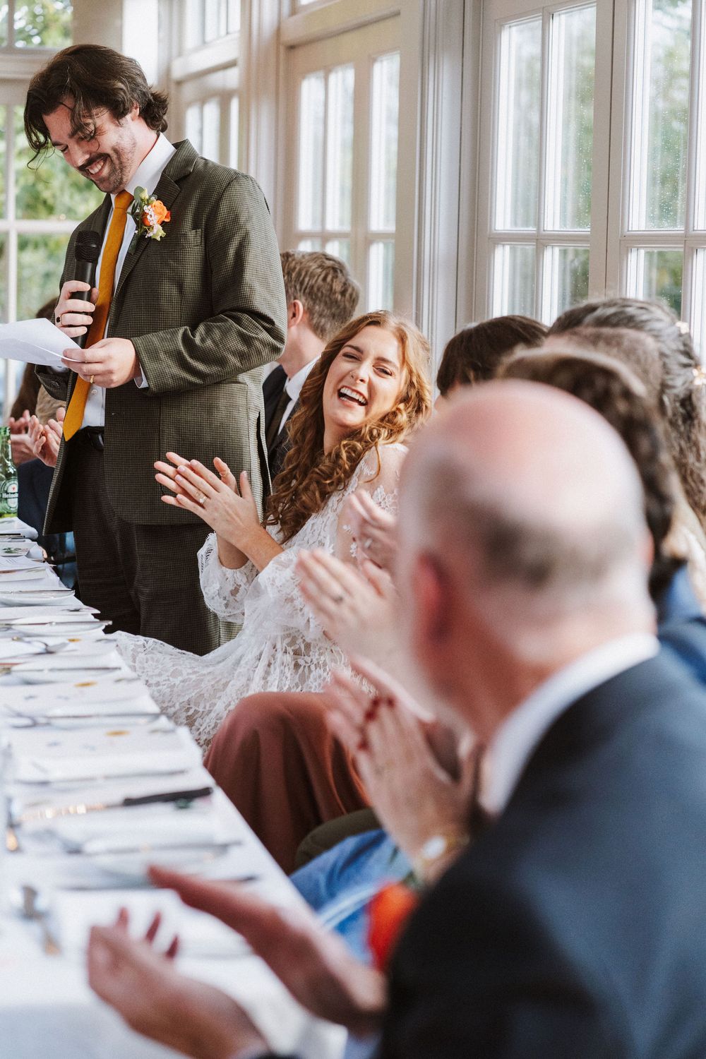 Wedding guests applaud and react emotionally during a reception speech in a bright sunlit venue.