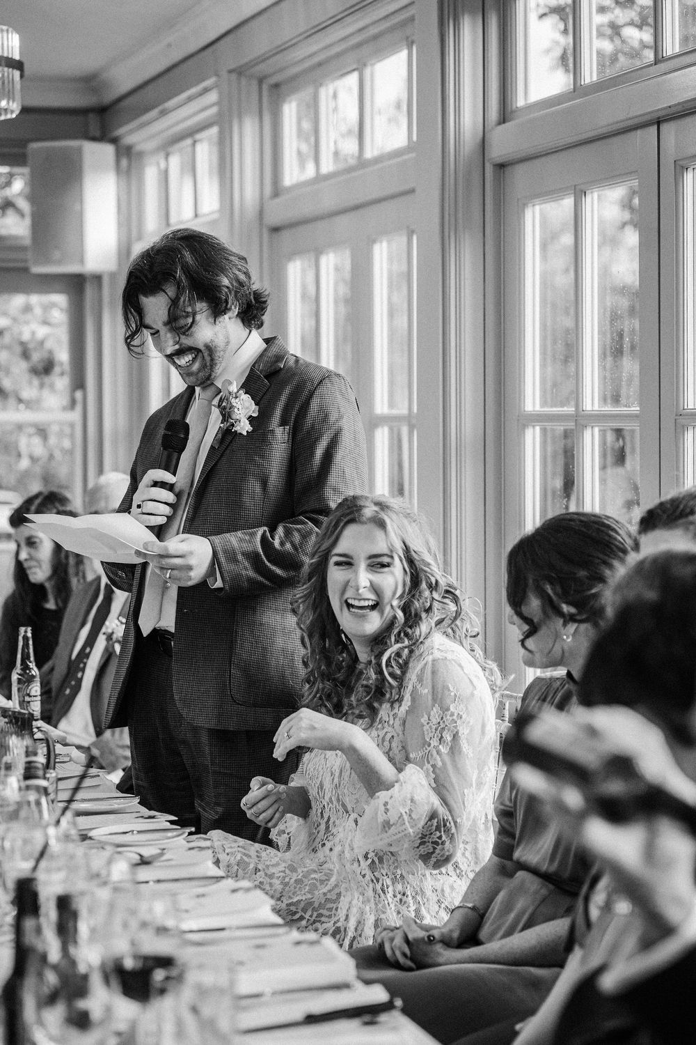 A black and white photo of a lively wedding reception with guests seated at a long table sharing laughs and drinks.
