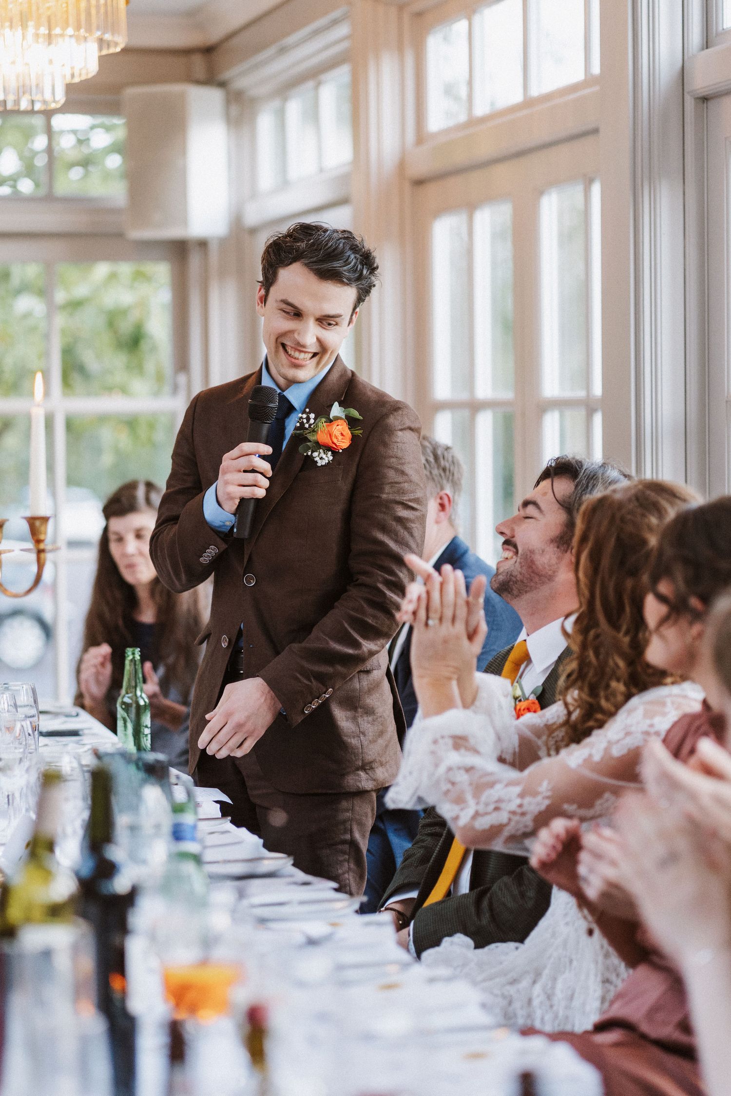 Wedding guests laugh and enjoy themselves during a reception dinner in an elegant sunlit venue.