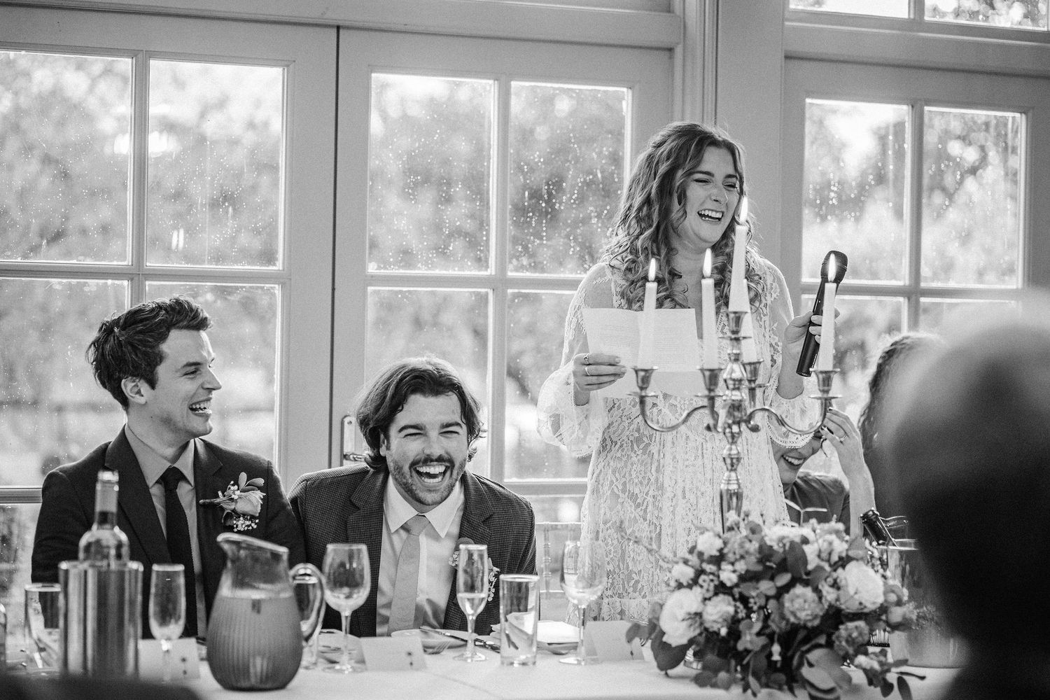 Wedding reception guests share a joyful moment of laughter during dinner at an elegant venue with large windows.