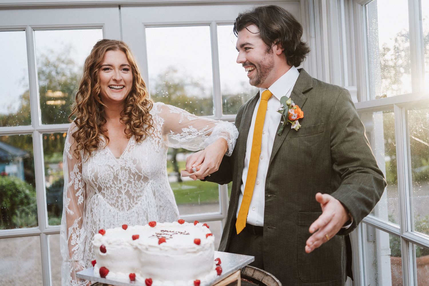 A couple shares a joyful moment while cutting their wedding cake in a bright sunlit room.