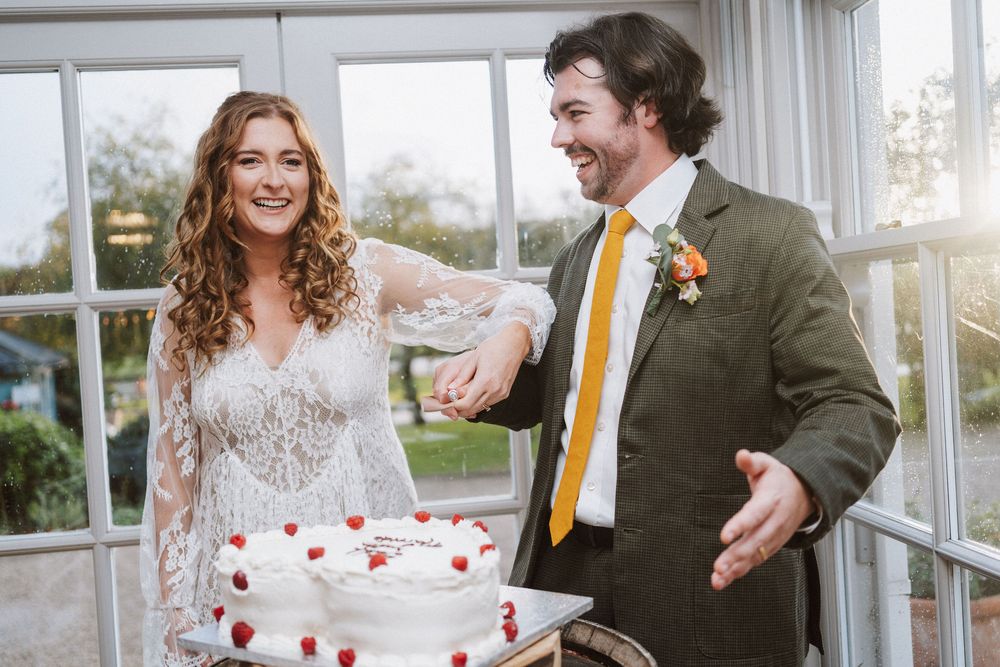 A couple shares a joyful moment while cutting their wedding cake in a bright sunlit room.