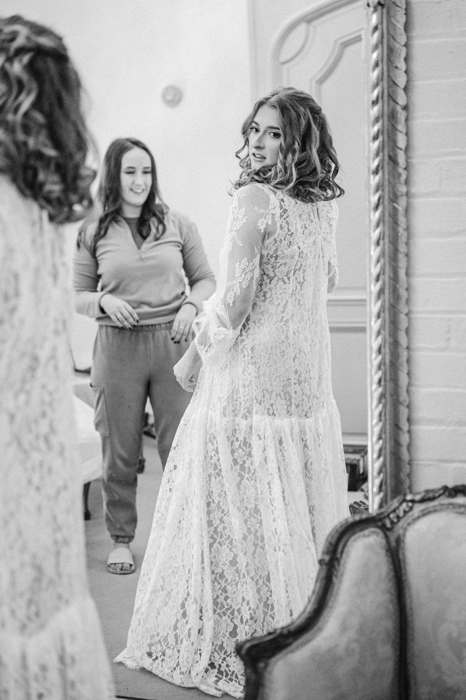 A toddler in a white lace dress twirls and plays in front of a decorative mirror in black and white photography.