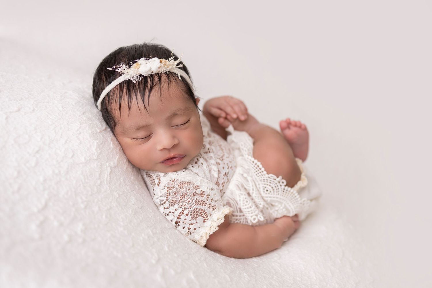 Newborn baby sleeping peacefully on soft white lace blanket in natural light.