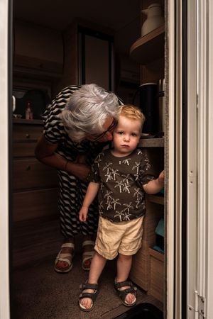 Small child peeks through sliding glass doors while wearing brown and beige outfit and sandals.